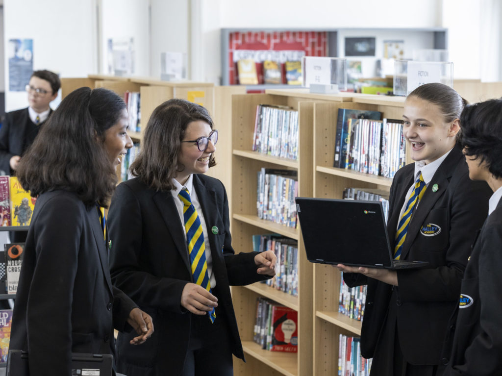 A small group of students are pictured conversing with each other in the Library area in the academy building. One is seen carrying a Chromebook laptop in her hands as she speaks.