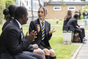 A small group of students are pictured sitting together on the school grounds, smiling and having a discussion.