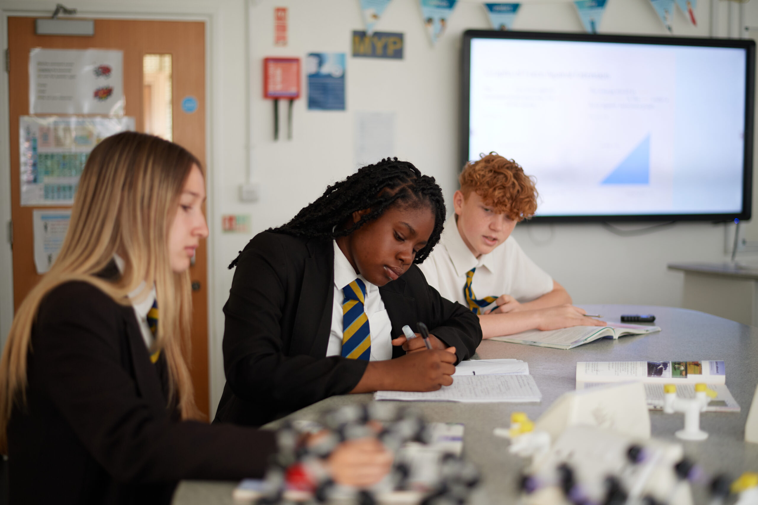 Three students sitting at a desk in a classroom, writing in workbooks