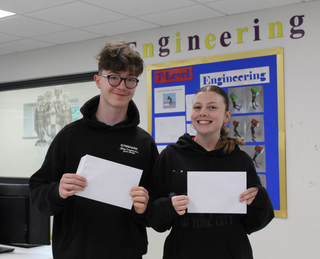 A young man and a young woman are standing together, both holding white envelopes or papers. They are smiling and appear to be in a classroom or similar setting, with a sign that says "Engineering" behind them