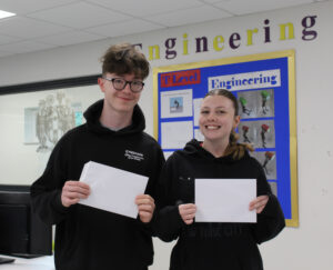 A young man and a young woman are standing together, both holding white envelopes or papers. They are smiling and appear to be in a classroom or similar setting, with a sign that says "Engineering" behind them